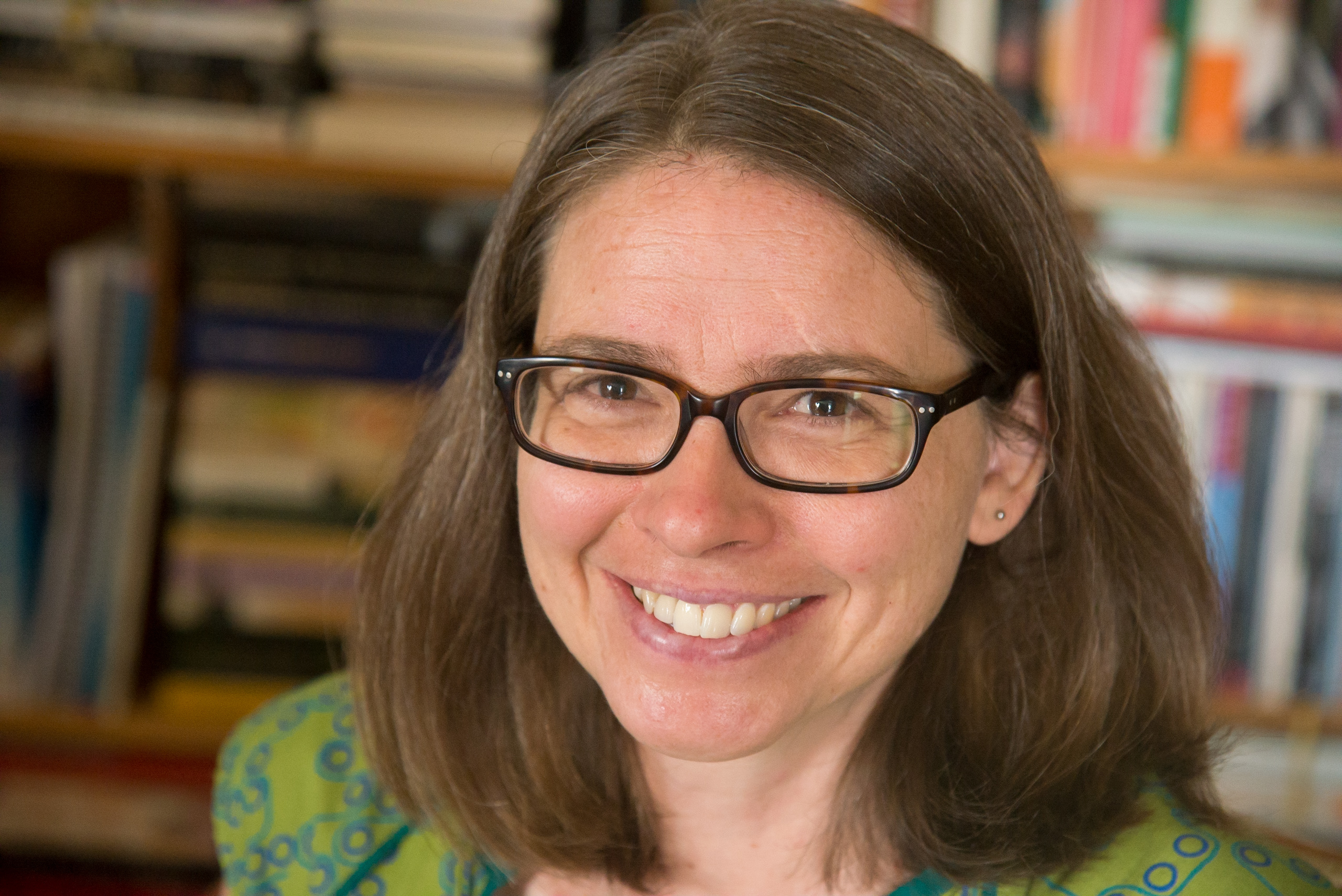 A white woman with shoulder-length brown hair and glasses smiles at the camera. Behind her are books on bookshelves.
