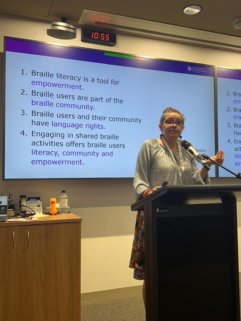 Jodie Martire giving a presentation at the SPEVI 2025 Conference in Brisbane. The slide behind her reads: 1. Braille literacy is a tool for empowerment. 2. Braille users are part of the braille community. 3. Braille users and their community have language rights. 4. Engaging in shared braille activities offers braille users literacy, community and empowerment.